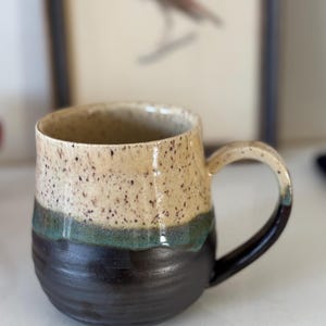 May include: Handmade ceramic mug with a speckled beige top, a teal band, and a dark brown base. The mug has a rounded shape and a curved handle. The background includes a framed bird illustration.