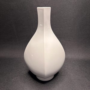 Könnte beinhalten: Eine weiße Keramikvase mit geometrischem Design. Die Vase hat einen breiten, abgerundeten Boden, der sich zu einem schmalen Hals und einer quadratischen Öffnung verjüngt. Die Oberfläche ist glatt und matt. Die Vase steht vor einem dunklen Hintergrund.