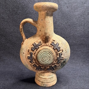 Könnte beinhalten: Eine beige Keramikvase mit Henkel und dekorativem kreisförmigem Design. Die Vase hat einen bauchigen Körper, einen schmalen Hals und einen Sockel. Das kreisförmige Design weist eine spiralförmige Mitte und ein dunkelbraunes zahnradartiges Muster auf.