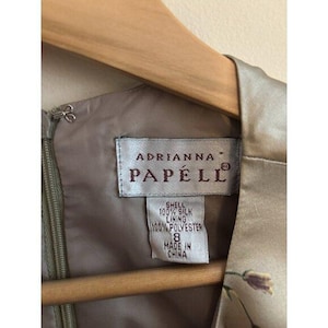 May include: Close-up of an Adrianna Papell dress tag. The label states "Shell 100% Silk, Lining 100% Polyester, 8, Made in China." The dress is a light tan colour and is on a wooden hanger.