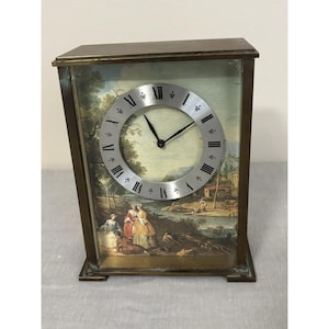 May include: A rectangular antique clock with a landscape scene on the face. The clock has a silver-colored circular clock face with black Roman numerals and hands. The frame is a dark bronze color.