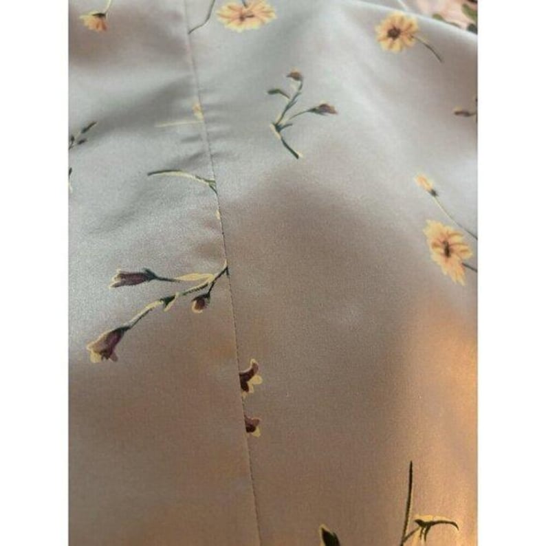 May include: Close-up of a pale grey fabric with a floral design. The pattern includes small yellow flowers and brown buds spread across the material. The fabric looks smooth, possibly silk or satin.