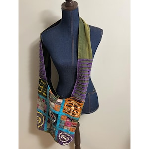 May include: A hobo bag featuring a patchwork design with various colorful fabric squares, including spiral and geometric patterns. The bag has a wide shoulder strap with horizontal purple and green fabric strips and a bright blue border.