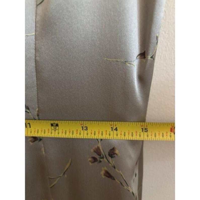 May include: Close-up of a light olive green fabric with a floral pattern. A yellow measuring tape shows a measurement of approximately 33 to 38 cm. The fabric has a subtle sheen.