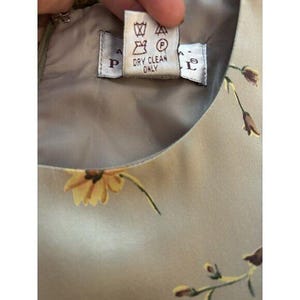 May include: Close-up of a dress with a floral pattern. The dress is a light beige colour with yellow and brown flowers. A tag reads "DRY CLEAN ONLY" with care symbols.