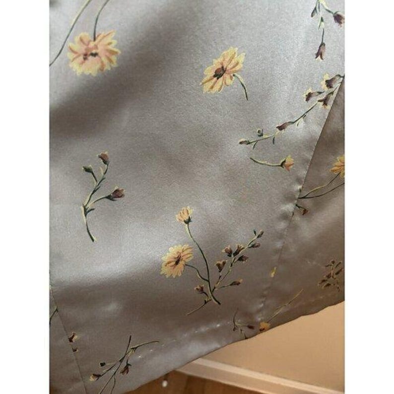 May include: A close-up of a light grey fabric with a floral design. The pattern includes yellow flowers and brown stems and buds. The fabric has a satin-like sheen, possibly part of a garment or home decor item.