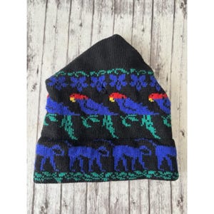May include: Black knit beanie with a colorful pattern. The design features blue parrots with red beaks and green leaves, along with blue animal figures. The hat is made of a soft, knitted material.