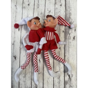 May include: Two plush elf dolls with vinyl faces and blue eyes. One elf wears a white hat with a red pom-pom, a red top, and white shorts with red checkered legs. The other elf wears a red and white striped hat, a red and white striped top, red shorts, and red and white striped legs.
