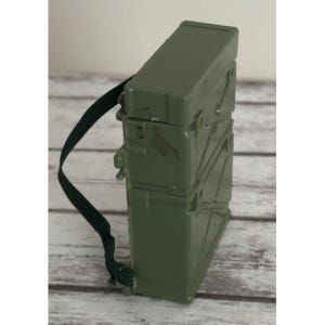 May include: An olive green rectangular box with a black strap. The box has a textured surface and appears to be made of plastic. The strap is attached to the box and is designed to be worn over the shoulder.