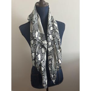 May include: A black and white snakeskin print scarf draped over a denim mannequin. The scarf has a square shape with a mix of black and white patterns. The scarf is made of a lightweight fabric.