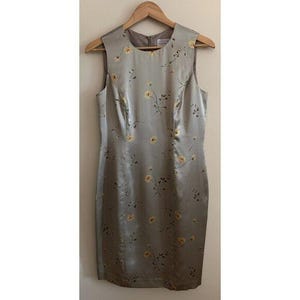 May include: A sleeveless, knee-length dress in a silvery-grey colour, decorated with a delicate floral pattern of yellow blossoms and slender green stems. The dress hangs on a wooden hanger, showing its tailored fit and subtle sheen.