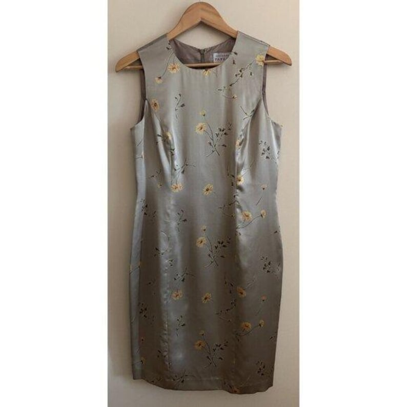 May include: A sleeveless, knee-length dress in a silvery-grey colour, decorated with a delicate floral pattern of yellow blossoms and slender green stems. The dress hangs on a wooden hanger, showing its tailored fit and subtle sheen.