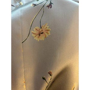 May include: Close-up of a pale grey fabric with a floral design. The pattern includes yellow and orange flowers with green stems and leaves. The fabric has a smooth, satin-like texture.