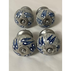 May include: A set of four round ceramic knobs featuring a crackled gray finish with blue and white teardrop-shaped patterns. Each knob has a silver-toned metal base and a central silver-toned metal decorative plate with a ridged edge.