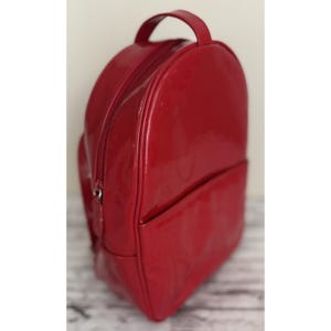 May include: A glossy red backpack with a top handle and a front pocket. The backpack has a zipper closure and a rounded shape. The material appears to be patent leather or a similar shiny material. The bag is sitting on a light-colored surface.