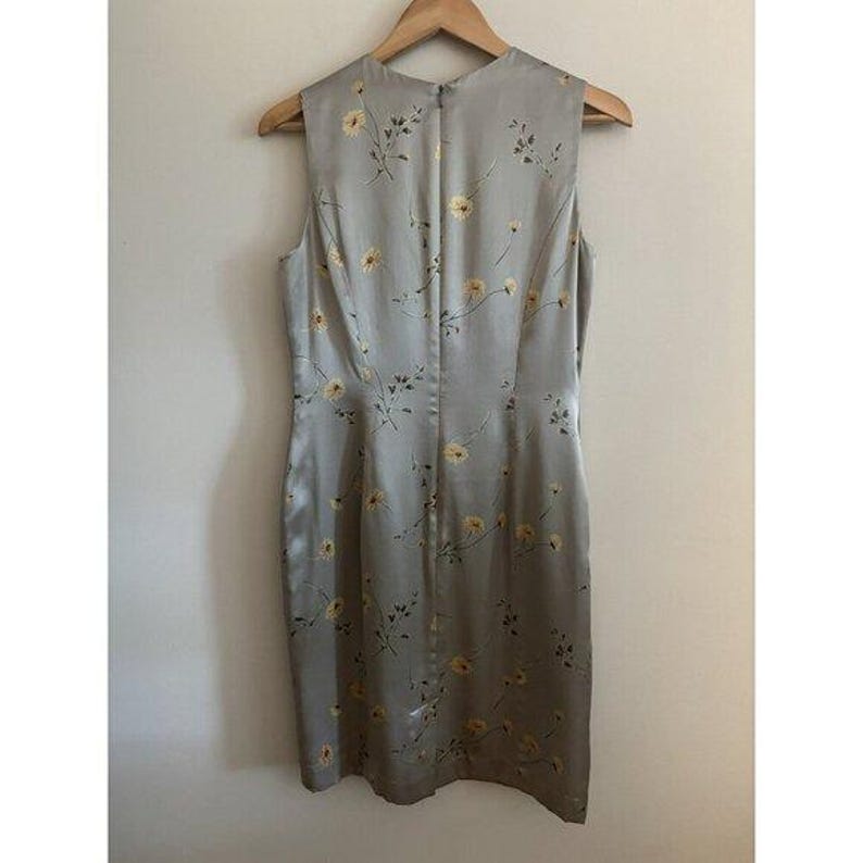 May include: A sleeveless, knee-length dress in a pale grey hue, decorated with a subtle yellow floral design. The dress is crafted from a silky fabric and is displayed on a wooden hanger. The back of the dress has a zip closure.