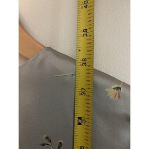 May include: A gray fabric with floral embroidery is measured with a yellow tape measure. The tape measure shows a length of 94 to 102 cm. The fabric is hanging on a wooden hanger.