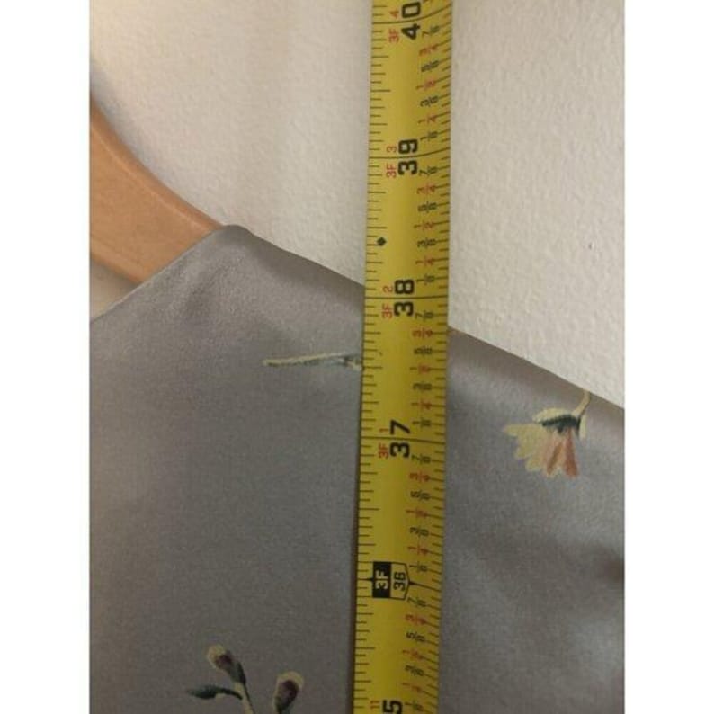 May include: A gray fabric with floral embroidery is measured with a yellow tape measure. The tape measure shows a length of 94 to 102 cm. The fabric is hanging on a wooden hanger.