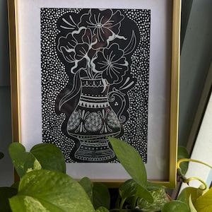 May include: A framed black and white art print featuring a vase with flowers. The artwork is in a gold frame and has a dotted background. The vase and flowers are detailed with white lines. The image is set against a light blue wall and surrounded by green plants.