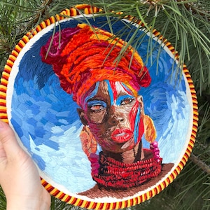 Puede incluir: Obra de arte bordada de una mujer con piel oscura que lleva un turbante naranja vibrante, labios rojos y pintura facial colorida. La obra de arte está enmarcada en un aro redondo con un borde amarillo, rojo y blanco. El fondo es un cielo azul.