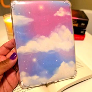 Puede incluir: Una funda protectora transparente para un dispositivo electrónico con un diseño de cielo. La funda tiene un paisaje de nubes rosa, azul y blanco con estrellas. La funda se sostiene sobre un libro abierto.