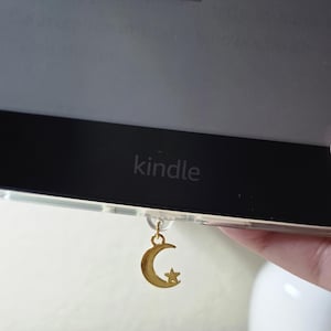 Op de afbeelding: Een goudkleurige maan- en sterrenbedel bevestigd aan een Kindle e-reader. De e-reader is zwart met het woord "kindle" erop gedrukt. De boektitel "jennifer jabaley" is bovenaan zichtbaar.