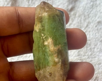 May include: A raw, natural green crystal with a rough, textured surface. The crystal has a light green hue with areas of tan and white. The crystal is held in a hand, showcasing its natural form and color variations. The crystal is a mineral specimen.