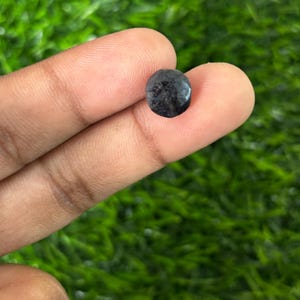 May include: A small, dark grey gemstone with a faceted surface. The gem is round and held between fingers against a blurred green background. The stone appears polished and reflects light.