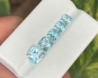 May include: Five light blue, square-cut gemstones are arranged diagonally on a white surface. The gems have a faceted appearance, reflecting light. The gemstones are likely aquamarine, a popular choice for jewelry.