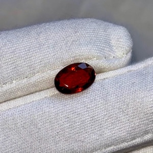 Könnte beinhalten: Ein facettierter, ovalförmiger, tiefroter Edelstein. Der Stein liegt auf einem weißen, strukturierten Stoff. Die polierte Oberfläche des Steins reflektiert das Licht und zeigt seine satte Farbe und innere Struktur. Geeignet für Schmuck.