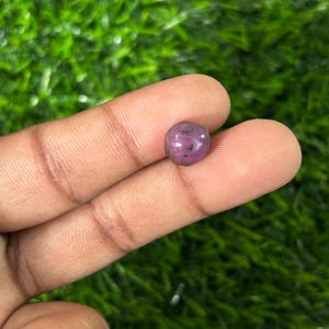 May include: A small, round, polished gemstone, likely a ruby, held between fingers. The gem is a deep, mottled pinkish-red color. The background is a blurred green, suggesting a natural setting.