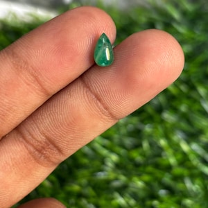 以下が含まれることがあります： 小さな涙の形をしたエメラルドの宝石。宝石は鮮やかな緑色で、磨かれているように見えます。エメラルドは、ぼやけた緑色の背景に対して、2本の指の間に保持されています。