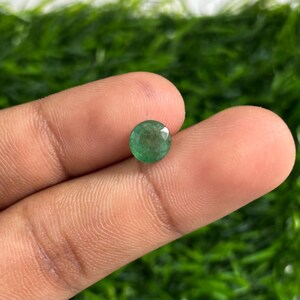 Puede incluir: Una piedra preciosa de esmeralda redonda y facetada. La gema es de color verde vibrante y se sostiene entre dos dedos sobre un fondo verde borroso. La esmeralda es una piedra preciosa que se usa a menudo en joyería.