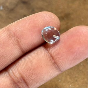 May include: A clear, oval-shaped gemstone with a slight blue tint. The gem is held between fingers, showcasing its polished surface and internal clarity. The gemstone is likely for jewelry making.