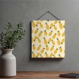 Op de afbeelding: Een vierkant kunstwerk met een herhaald patroon van gele ananassen met groene toppen op een crèmekleurige achtergrond. De kunst hangt aan een grijze muur. Een vaas met groene planten en een boek staan op een houten oppervlak.