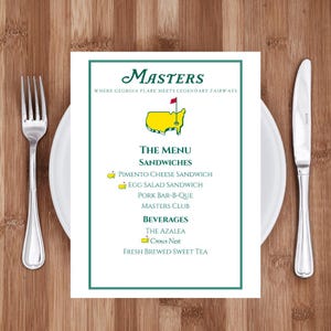 May include: A menu for the Masters golf tournament, featuring a green border and the tournament logo. The menu lists sandwiches like Pimento Cheese and Egg Salad, and beverages such as The Azalea and Fresh Brewed Sweet Tea. A fork and knife are on either side.