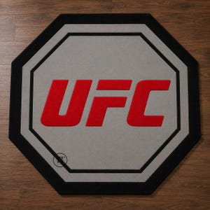 Peut inclure: Tapis gris de forme octogonale avec bordure noire et logo UFC rouge au centre. Le tapis présente un intérieur gris avec un contour noir autour du logo. Le tapis est posé sur un sol en bois.