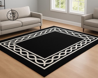 May include: A rectangular black rug with a white, repeating, intertwined design border. The rug is placed on a light-colored wooden floor, with a sofa and armchair in the background. The rug is a decorative home furnishing.
