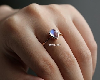 Puede incluir: Anillo de oro rosa con una piedra lunar en forma de pera, con iridiscencia azul y púrpura. El anillo tiene una banda delgada. La imagen incluye el texto "Silver Lush".