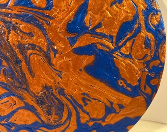 Puede incluir: Obra de arte circular con un patrón arremolinado de pintura naranja y azul. Los colores se mezclan y crean un efecto marmolado, con el azul pareciendo fluir a través del naranja.