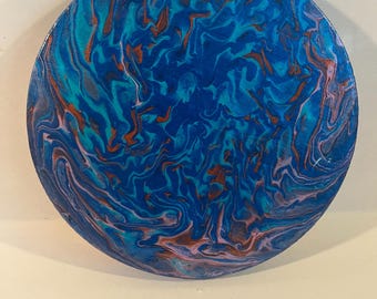 Puede incluir: Obra de arte circular y abstracta con un diseño fluido. La pieza presenta un patrón arremolinado de azules, turquesas, naranjas y rosas. Este artículo decorativo es probablemente para exhibir en la pared o sobre una mesa, con un diámetro de aproximadamente 25 cm.