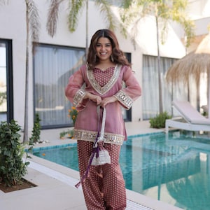 Könnte beinhalten: Eine Frau in einem traditionellen, mauvefarbenen Outfit mit goldenen und silbernen Verzierungen. Das Outfit besteht aus einer Tunika und einer Hose mit Muster. Sie hält eine kleine, silberne Kordeltasche. Im Hintergrund ein Pool und Palmen.