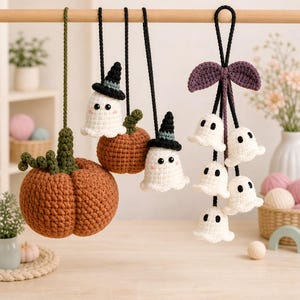 May include: A collection of crocheted hanging ornaments featuring two orange pumpkins with green stems, two white ghosts wearing black witch hats, and a cluster of five small white ghosts suspended from a purple crocheted bow.