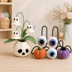 May include: A crochet set featuring a white skull-shaped pot with green stems and three white ghost-shaped flowers, alongside three round crochet eyeballs with colorful irises and two small crochet pumpkins in orange and purple colors.