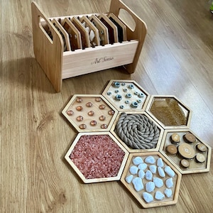 Puede incluir: Una caja de almacenamiento de madera con la etiqueta Art Senso que contiene varios tableros sensoriales hexagonales. Los tableros presentan diversos materiales táctiles, incluyendo tuercas metálicas, cuentas de vidrio ámbar, purpurina dorada, una cuerda enrollada, cristales rosas, piedras blancas y rodajas de madera.