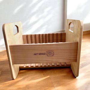 Könnte beinhalten: Bücherregal aus Holz für Kinder mit natürlichem Finish. Das Regal hat ein rechteckiges Design mit Lattenrücken und -boden sowie eine durchgehende Frontplatte mit dem Text "ART SENSO" und einem Handlogo. Die Seiten haben bogenförmige Ausschnitte.