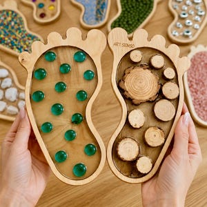 Könnte beinhalten: Zwei fußförmige sensorische Holzbretter mit heller Holzoberfläche. Ein Brett enthält eingebettete, durchscheinende grüne Glassteine, während das andere verschiedene kreisförmige Holzscheiben unterschiedlicher Größe aufweist. Der Text ART SENSO ist eingraviert.