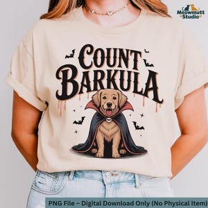 May include: Beige t-shirt featuring a cartoon illustration of a golden retriever dressed as a vampire, with the text "COUNT BARKULA". The design includes bats and dripping blood accents, perfect for Halloween.