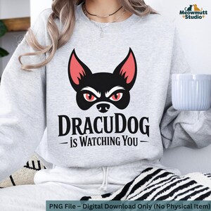 May include: A light gray sweatshirt featuring a black dog face with red eyes and ears. The text "DRACUDOG is WATCHING YOU" is printed below the graphic. The sweatshirt is part of the Meowmutt Studio collection.