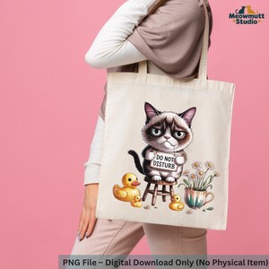 May include: A beige tote bag featuring a grumpy cat illustration holding a "DO NOT DISTURB" sign. The design includes yellow rubber ducks, daisies, and a teacup. The bag is held by a person against a pink background.